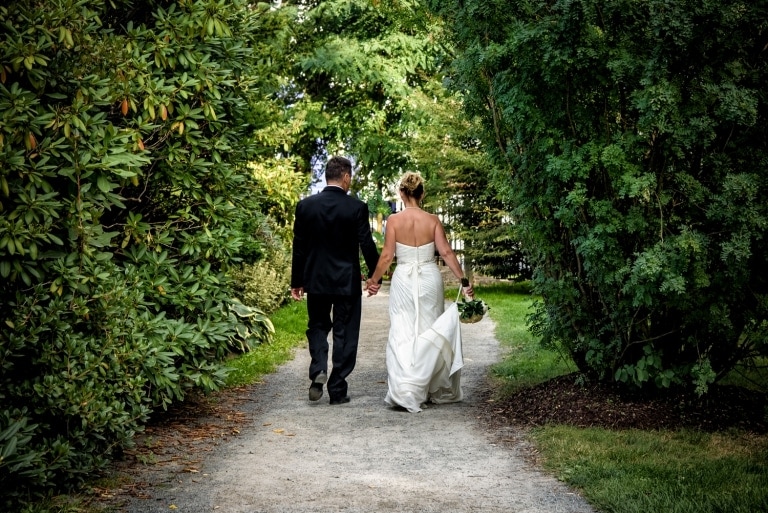 The bride and groom wedding photography in the Halifax public gardens walking along a pathway among the evergreen trees.