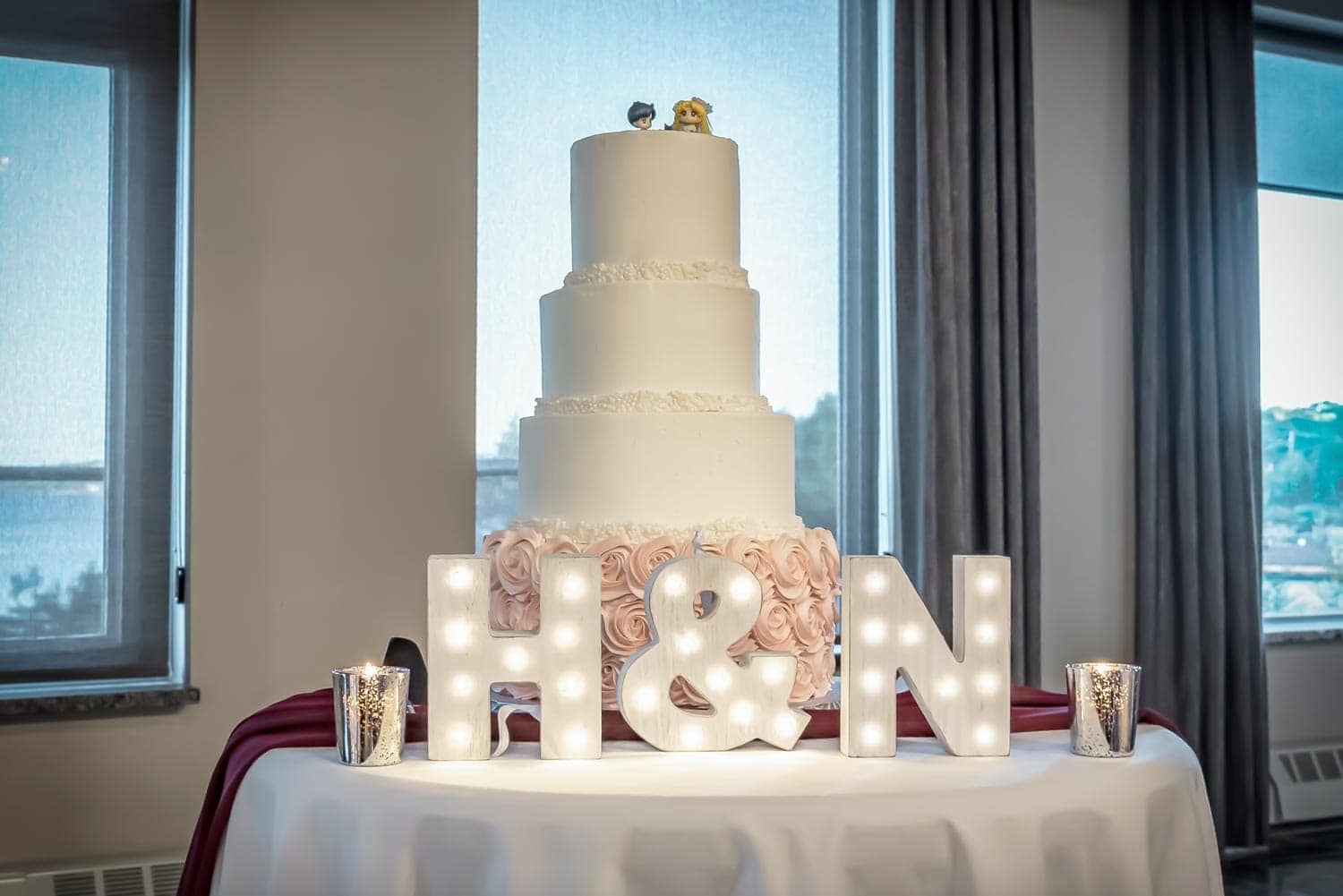 A 4 tier wedding cake with figurine wedding cake toppers and marquee letters lit up at the Bedford Basin Market.