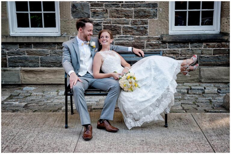 Bride and groom sitting on a bench outdoors, smiling, with the bride holding a bouquet, showcasing their wedding day in Halifax.