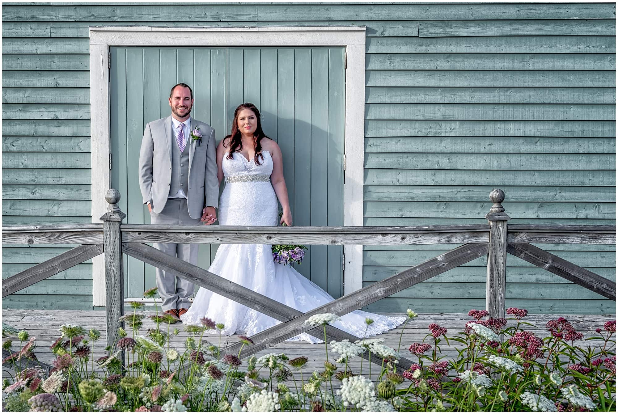 The bride and groom pose for wedding photos at their White Point wedding against a rustic barn style building.