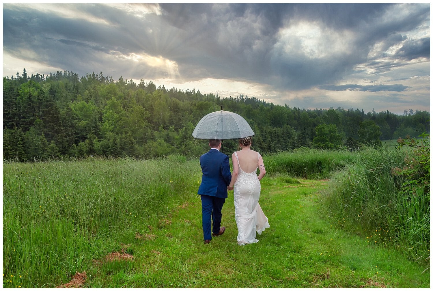 The bride and groom walk hand in hand through a field in the rain on their wedding day at Parkers Barn in Hopewell NS.