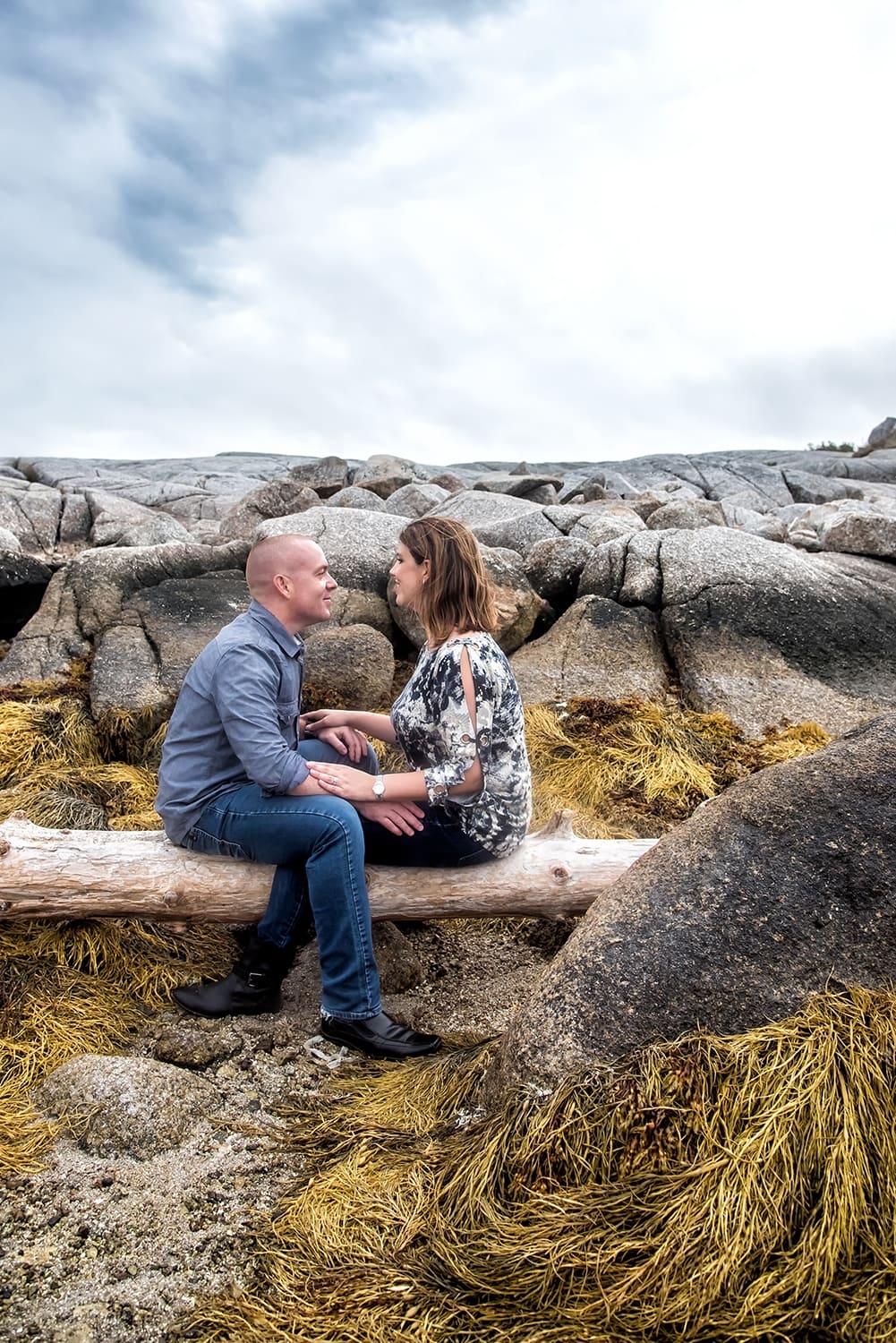 A love couple face each other holding hands sitting on a log at Peggys Cove for their engagement photos.
