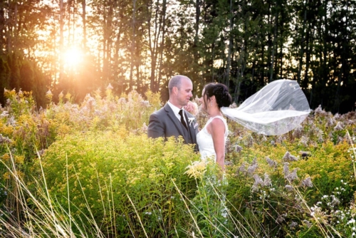 The bride and groom standing in a field at sunset with her veil flying behind her at the Old Orchard Inn Wolfville, NS.